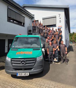 Bild Firmengebäude mit dem gesamten Team auf der Treppe und danneben steht ein Firmenwagen.