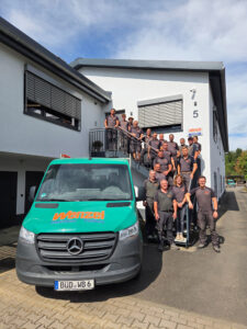 Bild mit dem gesamten Team der Firma Wenzel-Bau auf einer Treppe des Gebäudes. Firmenwagen in grün ist auch zu sehen.